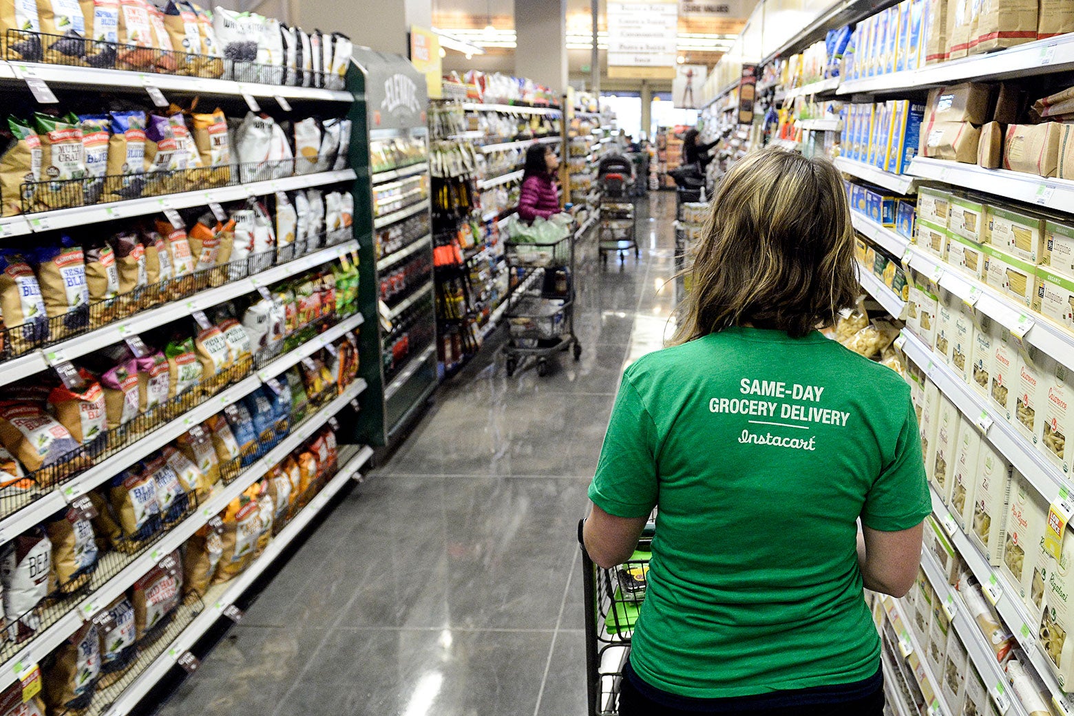 A woman in a green Instacart shirt is seen from the back pushing a cart down a grocery store aisle. 