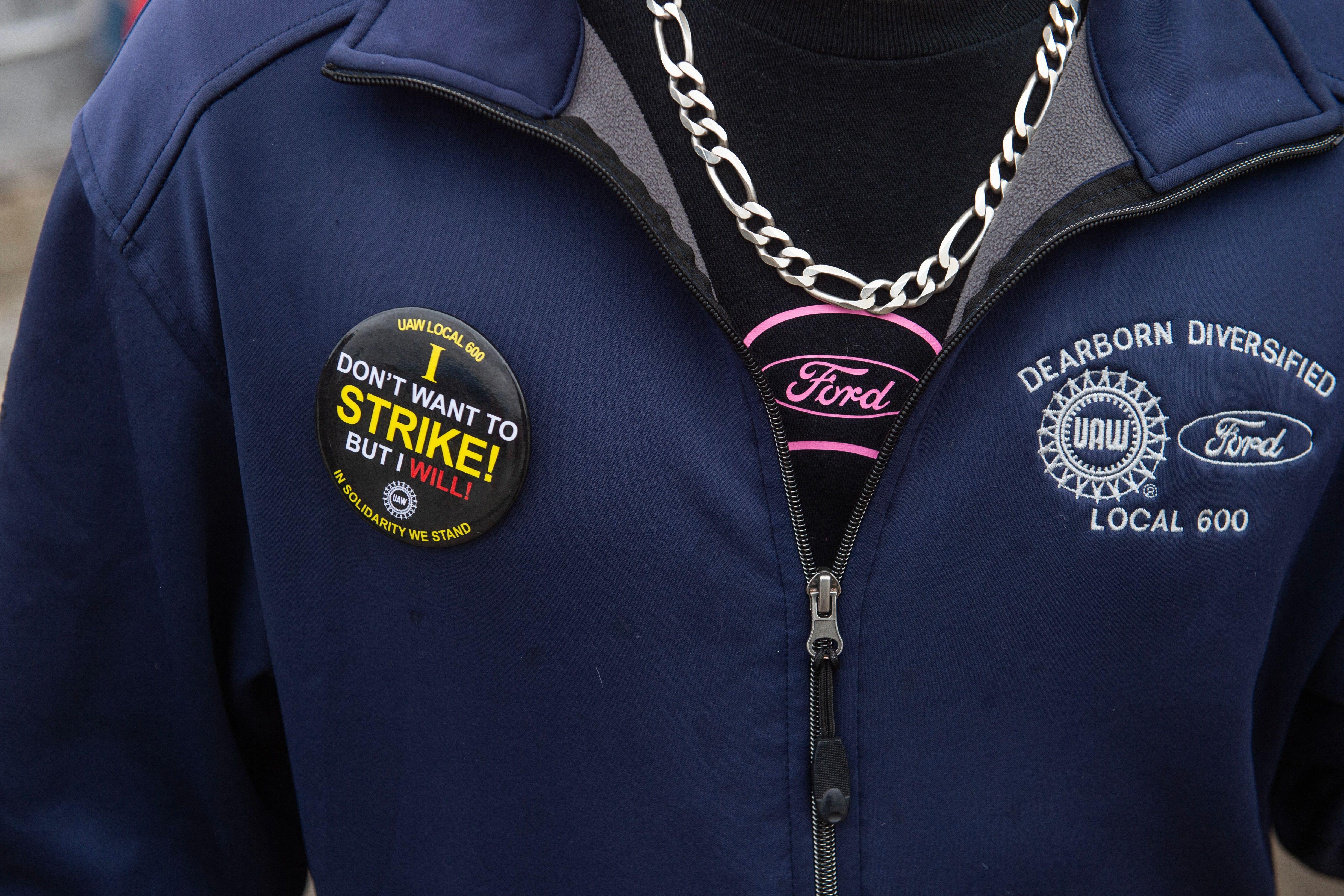 A close-up image of the chest of the worker on the UAW picket line, focused on a pin that reads: "I DON'T WANT TO STRIKE! BUT I WILL!" The person is wearing a silver chain necklace, and a front-zipped blue sweatshirt with emblems that read: "DEARBORN DIVERSIFIED, UAW, FORD, LOCAL 600." A black T-shirt with a pink Ford logo can be seen peeking out underneath.